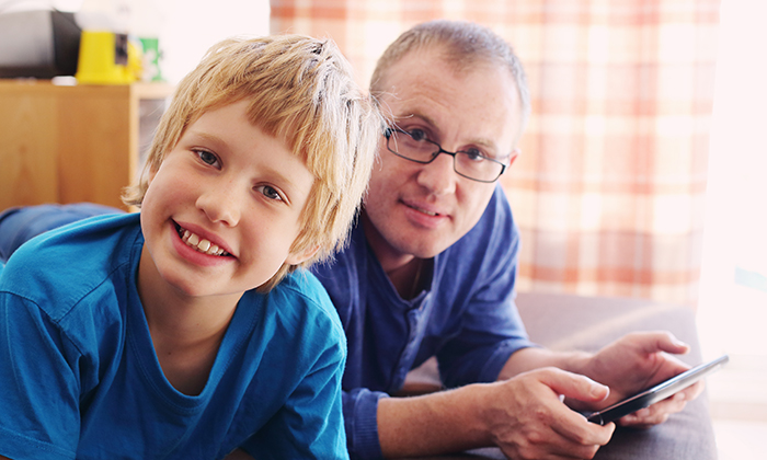A man and a young boy are smiling at the camera. The man is holding a tablet in his hands.